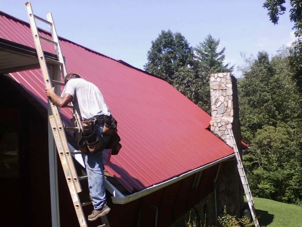 Roofer on a ladder beside a red metal roof and stone chimney — Soffit & Fascia Repair by Roofing Brothers in Commerce Township